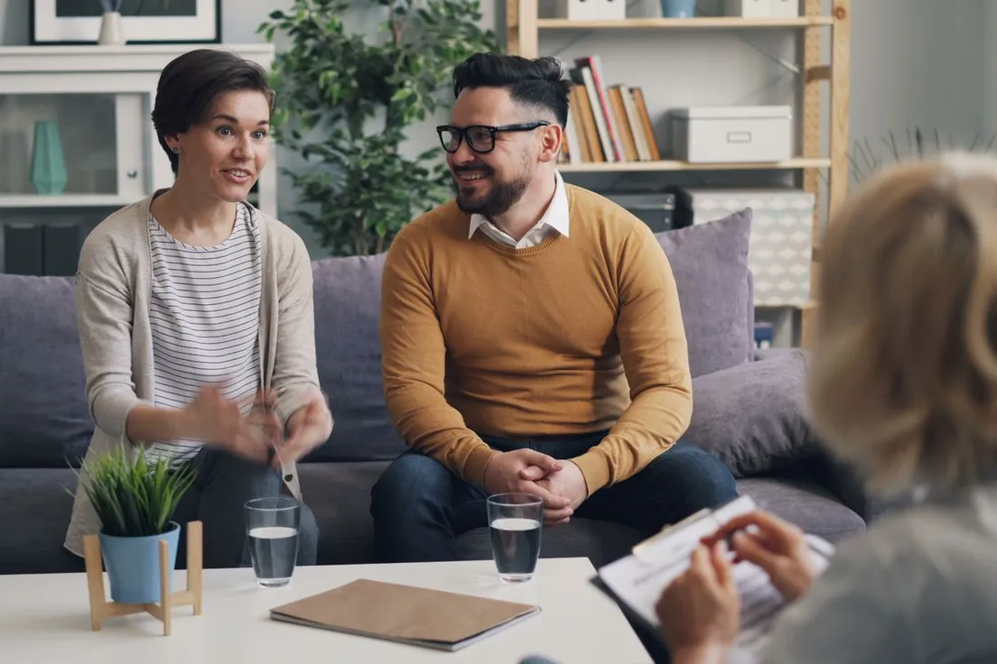 A smiling couple talking with a therapist in a cozy office with bookshelves and plants